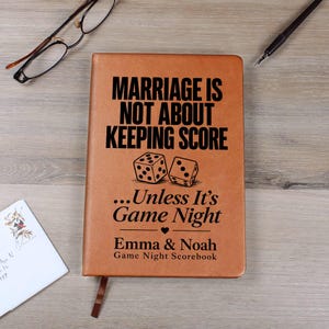 May include: A tan leather-bound notebook featuring the black text "MARRIAGE IS NOT ABOUT KEEPING SCORE...Unless It's Game Night" with a small heart icon, two dice illustrations, and the names "Emma & Noah" printed on the front cover.