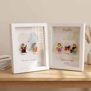 May include: Two white shadow box frames containing custom displays. The left frame features a heart-shaped map, two minifigures labeled Michael and Maria, and the text The place where we said I do forever. The right frame shows a house, three minifigures labeled Emily, Teddy, and Josh, and the text Our first home and Cheers to a new chapter.
