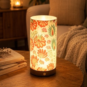 May include: A cylindrical table lamp featuring a white shade with a repeating pattern of orange, red, and green floral and leaf motifs. The lamp has a solid dark base with a small circular power button on the front, emitting a warm, soft glow.