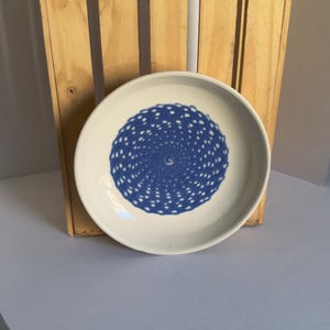 May include: A round, off-white ceramic bowl featuring a centered, circular pattern of small, dark blue dots arranged in a swirling, spiral design that radiates outward from the center of the bowl's interior base.