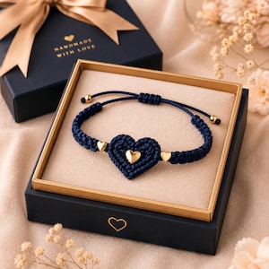 May include: A dark blue macrame bracelet featuring a central crocheted heart, accented with three small gold-toned heart charms and adjustable sliding knots, presented in a black gift box with the text "HANDMADE WITH LOVE" and a gold ribbon.