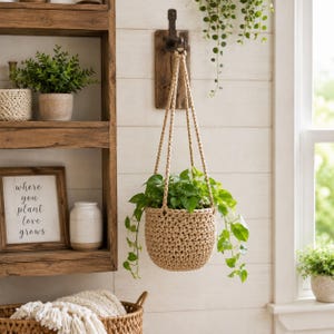 Puede incluir: Una cesta colgante de ganchillo en color beige, hecha de cuerda gruesa y suspendida por tres cuerdas trenzadas a juego desde un gancho de pared de metal oscuro. La cesta contiene una planta potos verde con enredaderas colgantes.