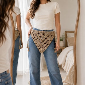 May include: A beige triangular crochet hip scarf featuring an openwork diamond pattern and scalloped edges. The accessory is tied at the sides with thin cords finished with small wooden beads and tassels, worn over blue denim jeans and a white t-shirt.