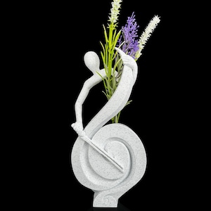 Könnte beinhalten: Eine weiße, gesprenkelte Skulptur in Form eines Violinschlüssels mit einer stilisierten menschlichen Figur, die in die Kurve integriert ist. Die Skulptur hält ein Bündel künstlicher Blumen mit grünen, weißen und violetten Stielen in ihrer Mitte.