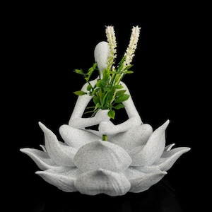Pode incluir: Uma estatueta branca salpicada de uma pessoa sentada na posição de lótus sobre uma base de flor de lótus grande com várias pétalas. A figura segura um pequeno ramo de folhas verdes e dois caules florais brancos, finos e texturizados.