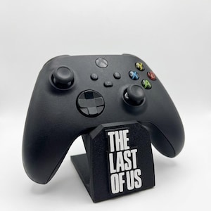 Pode incluir: Um comando de videojogos sem fios preto pousado num suporte preto. O suporte apresenta o texto branco "THE LAST OF US" impresso na parte frontal. O comando inclui dois joysticks analógicos, um D-pad e quatro botões de ação coloridos.