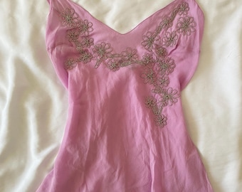 May include: A light pink camisole featuring a V-neckline and thin spaghetti straps. The garment is adorned with intricate, dark-toned floral embroidery that cascades from the neckline down the left side, finished with a simple hemline.
