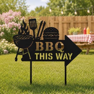 Op de afbeelding: Een zwart metalen pijl-vormig tuinbord met een uitgesneden ontwerp van een barbecue met vlammen, een spatel, een tang en een hamburger. Het bord toont de tekst "BBQ THIS WAY" in hoofdletters en wordt ondersteund door twee dunne metalen staven.