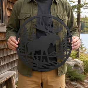 May include: A circular black metal wall art piece featuring a silhouette of a moose standing in a forest with pine trees and mountains in the background. The design is cut out from a single flat sheet of metal, creating a detailed nature scene.