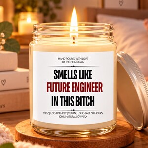 Pode incluir: Uma vela num frasco de vidro transparente com um pavio aceso e um rótulo branco com o texto: HAND POURED WITH LOVE BY THE NESTTORAA, SMELLS LIKE FUTURE ENGINEER IN THIS BITCH, 9 OZ | ECO-FRIENDLY | VEGAN | LONG LAST 50 HOURS 100% NATURAL SOY WAX.