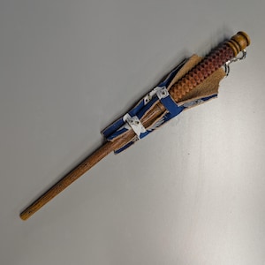 Puede incluir: Una varita de madera con un mango tallado y una funda de cuero y tela. La varita es marrón con un agarre texturizado, y la funda es azul con un estampado blanco. Un anillo de metal plateado está unido a la varita.
