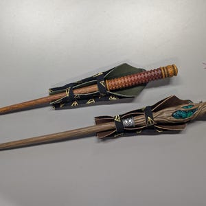 May include: Two wooden wands with decorative handles and protective sheaths. One wand has a brown and red diamond pattern, while the other has a wood-grain design with green accents. The sheaths are black with gold symbols.