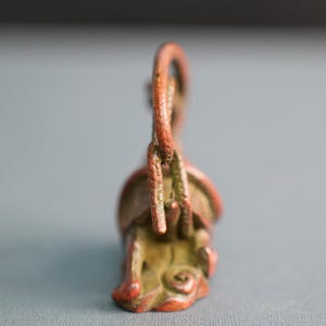 Puede incluir: Una peque&ntilde;a escultura de metal de color marr&oacute;n rojizo con un asa curva y texturizada que se arquea sobre un cuerpo central. La base muestra intrincados patrones en espiral y formas org&aacute;nicas, con un acabado de p&aacute;tina envejecida.
