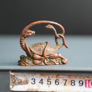 Puede incluir: Una figura de metal en tono bronce que representa una tortuga entrelazada con una serpiente. La tortuga tiene un caparaz&oacute;n texturizado y la serpiente se arquea sobre su cabeza. Una cinta m&eacute;trica muestra una anchura de unos 9 cm.