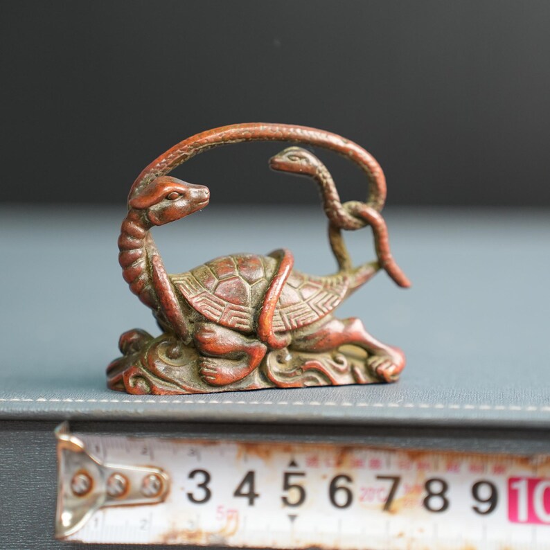 Puede incluir: Una figura de metal en tono bronce que representa una tortuga entrelazada con una serpiente. La tortuga tiene un caparaz&oacute;n texturizado y la serpiente se arquea sobre su cabeza. Una cinta m&eacute;trica muestra una anchura de unos 9 cm.