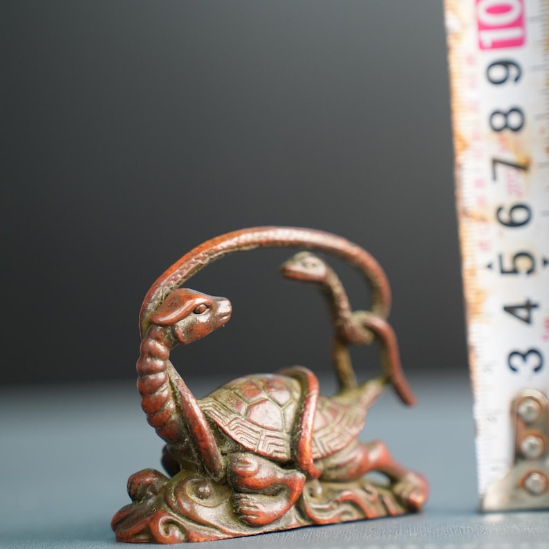 Puede incluir: Una escultura de metal en tono bronce de una tortuga entrelazada con una serpiente, colocada junto a una cinta m&eacute;trica que muestra una altura de aproximadamente 9 cm. La tortuga tiene un patr&oacute;n de caparaz&oacute;n detallado y la serpiente se curva.
