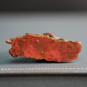 Puede incluir: Una peque&ntilde;a figura de bronce desgastada con una base texturizada pintada en rojo anaranjado. El objeto muestra una p&aacute;tina met&aacute;lica envejecida con signos visibles de desgaste y oxidaci&oacute;n, presentando una forma compleja e irregular con detalles esculpidos.