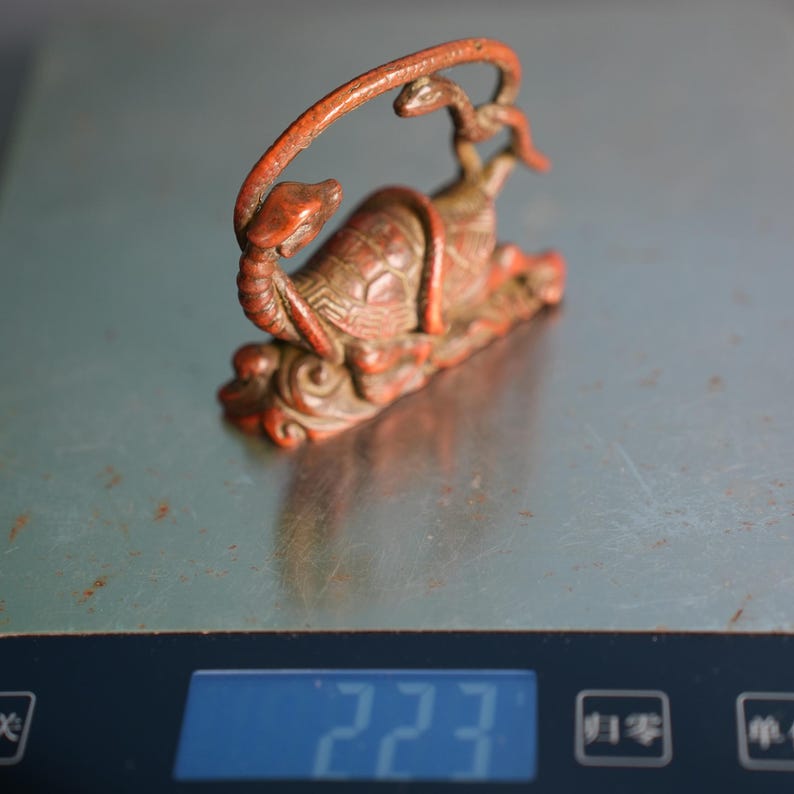 Puede incluir: Una figura tallada de color marr&oacute;n rojizo que representa una tortuga con una serpiente enroscada alrededor de su caparaz&oacute;n. El objeto est&aacute; colocado sobre una b&aacute;scula digital que muestra un peso de 223 gramos. La tortuga tiene patrones detallados.