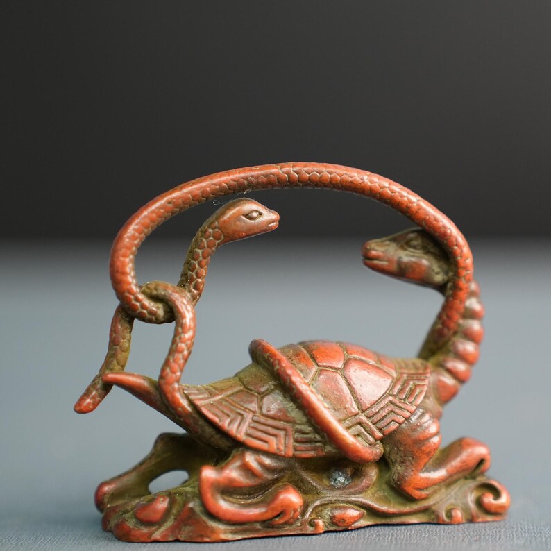 Puede incluir: Una figura tallada de color marr&oacute;n rojizo que representa una tortuga con un caparaz&oacute;n estampado y dos serpientes entrelazadas alrededor de su cuerpo. La escultura presenta texturas intrincadas en las escamas y el caparaz&oacute;n.