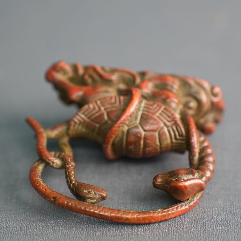 Puede incluir: Un netsuke tallado de color marr&oacute;n rojizo que presenta una tortuga con un patr&oacute;n de caparaz&oacute;n detallado entrelazada por una serpiente. La escultura muestra texturas intrincadas y un acabado desgastado, con formas animales compactas.