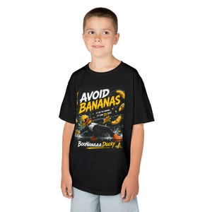 May include: A black short-sleeve t-shirt featuring a graphic print of a duck wearing sunglasses and a suit, sliding on a banana peel. The text reads AVOID BANANAS, IT'S NOT THE BANANA... IT'S HOW YOU DUCK, and BOONANAAA DUCKY with banana illustrations.