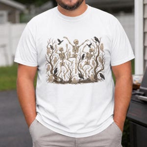May include: A white t-shirt featuring a detailed graphic print of three skeletons dancing among bare, gnarled trees. The illustration includes several black crows perched on branches and small, pale ghost figures scattered throughout the scene.