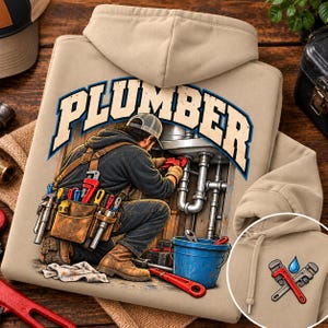 May include: A beige hooded sweatshirt featuring a graphic of a plumber working under a sink with various tools. The word PLUMBER is printed in large letters above the figure. A smaller graphic of crossed pipe wrenches and a blue water drop is visible.