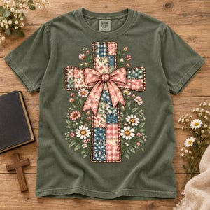 May include: An olive green short-sleeve t-shirt featuring a central graphic of a cross made of various patchwork patterns in blue, pink, and white. A large pink bow is tied around the centre of the cross, surrounded by illustrations of daisies.