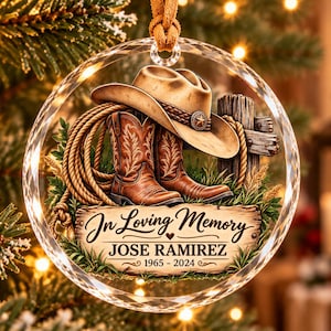 May include: A round, clear glass ornament featuring a western-themed illustration of brown cowboy boots, a tan cowboy hat, and a coiled rope. The text reads In Loving Memory, JOSE RAMIREZ, and 1965 - 2024, with a small heart icon.