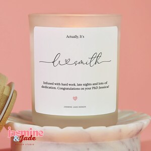 May include: A frosted glass candle with a white label featuring the text Actually, It's dr smith, Infused with hard work, late nights and lots of dedication. Congratulations on your PhD Jessica!, and a small pink heart icon by Jasmine Jade Design.