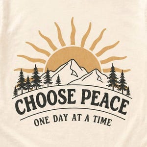 Puede incluir: Una camiseta de manga corta de color crema con un gráfico de un sol saliendo sobre montañas y pinos. El texto "CHOOSE PEACE" está impreso en letras negras grandes, con "ONE DAY AT A TIME" en letras negras más pequeñas debajo.
