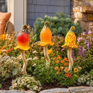 Puede incluir: Tres estacas de jardín de cerámica con forma de flores acampanadas o setas, cada una con un tallo largo, delgado y de color blanco roto. Las cabezas son brillantes y están coloreadas en tonos vibrantes de rojo anaranjado, naranja sólido y amarillo con motas rojas.