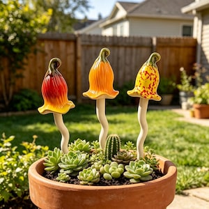 Puede incluir: Tres estacas de jardín de cerámica con forma de hongo, tallos blancos ondulados y sombreros en forma de campana de colores rojo, naranja y amarillo con motas rojas, plantadas en una maceta de terracota con suculentas y un cactus.