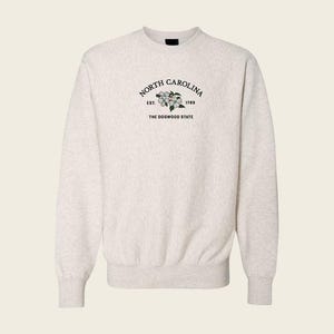 May include: A light heather gray crewneck sweatshirt featuring an embroidered design on the chest. The text reads NORTH CAROLINA, EST. 1789, and THE DOGWOOD STATE, surrounding a small illustration of white dogwood flowers with green leaves.