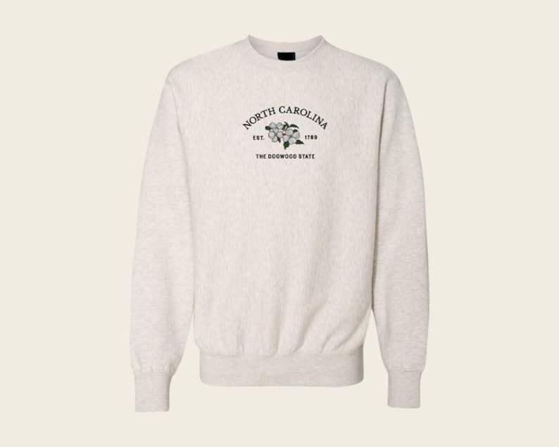 May include: A light heather gray crewneck sweatshirt featuring an embroidered design on the chest. The text reads NORTH CAROLINA, EST. 1789, and THE DOGWOOD STATE, surrounding a small illustration of white dogwood flowers with green leaves.