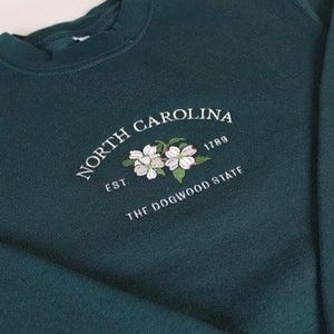 May include: A dark forest green crewneck sweatshirt featuring white embroidered text that reads NORTH CAROLINA EST 1789 THE DOGWOOD STATE, centered above an embroidered illustration of two white dogwood flowers with green leaves.