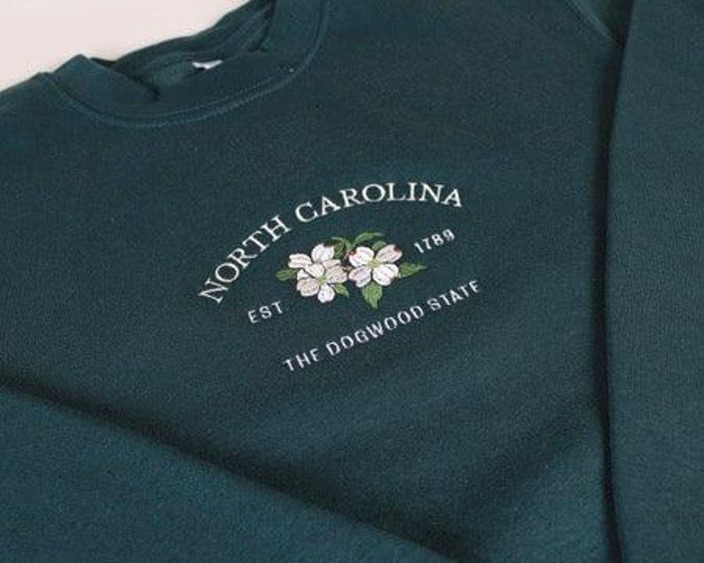 May include: A dark forest green crewneck sweatshirt featuring white embroidered text that reads NORTH CAROLINA EST 1789 THE DOGWOOD STATE, centered above an embroidered illustration of two white dogwood flowers with green leaves.