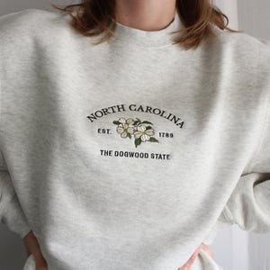 May include: A light gray heathered crewneck sweatshirt featuring dark green embroidered text that reads North Carolina, Est. 1789, and The Dogwood State, centered above an embroidered illustration of white dogwood flowers with green leaves.