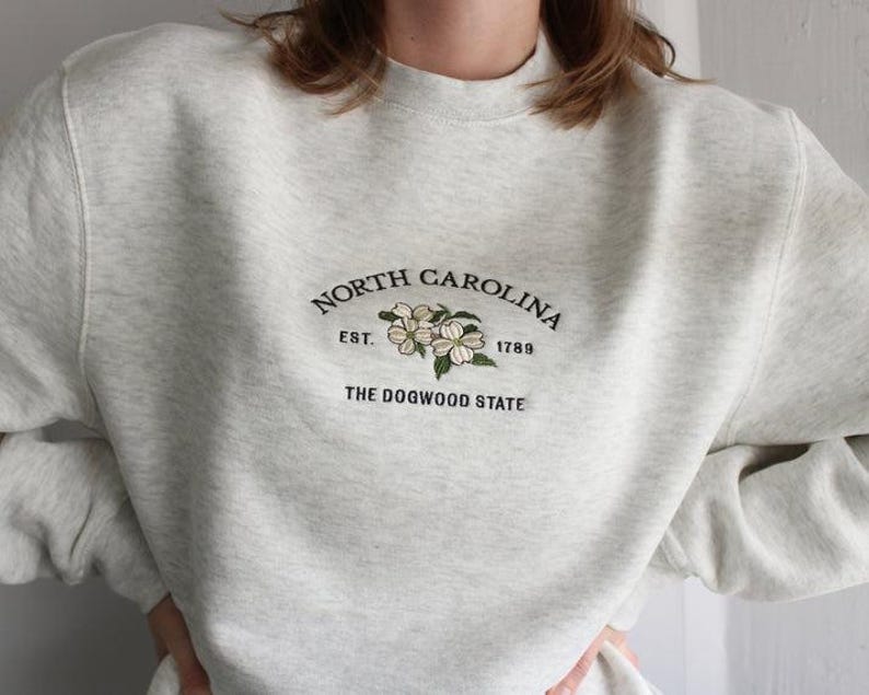 May include: A light gray heathered crewneck sweatshirt featuring dark green embroidered text that reads North Carolina, Est. 1789, and The Dogwood State, centered above an embroidered illustration of white dogwood flowers with green leaves.