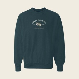 May include: A dark teal crewneck sweatshirt featuring white text that reads North Carolina, Est. 1789, and The Dogwood State, centered above a small illustration of white dogwood flowers with green leaves on the upper chest area.