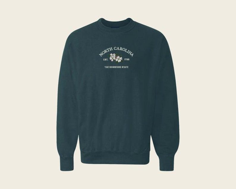 May include: A dark teal crewneck sweatshirt featuring white text that reads North Carolina, Est. 1789, and The Dogwood State, centered above a small illustration of white dogwood flowers with green leaves on the upper chest area.