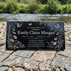 May include: A rectangular black memorial plaque featuring the text In Loving Memory Of Emily Claire Morgan, May 12, 1962 – August 24, 2025, and a poem. The plaque is decorated with colourful butterflies and detailed illustrations of wildflowers.