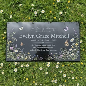 May include: A rectangular black memorial plaque featuring the text In Loving Memory, Evelyn Grace Mitchell, March 14, 1948 – May 21, 2025, and a tribute verse. The plaque is decorated with colourful butterflies and various wildflowers.