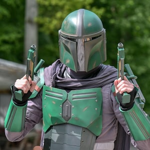 Könnte beinhalten: Eine Person in einem grünen und silbernen Mandalorianer-Helm und -Rüstung, die zwei grüne Blaster hält. Das Kostüm hat ein verwittertes Aussehen, mit einem grauen Schal und Handschuhen. Der Hintergrund ist verschwommen mit grünem Laub.