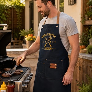Könnte beinhalten: Eine marineblaue Schürze mit braunen Lederriemen am Hals und an den Taschen, verziert mit dem gelben Sticktext 'GRILL MASTER' über einem gekreuzten Gabel- und Pfannenwender-Symbol sowie dem Namen 'AIDEN' mit Sternen und zwei großen Vordertaschen.