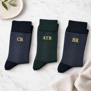 May include: Three pairs of socks featuring a herringbone pattern. Two pairs are dark navy blue with black cuffs and toes, embroidered with yellow initials CR and BR. The center pair is dark green with matching cuffs, toes, and initials ATB.