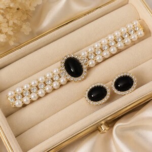 May include: A gold-toned bracelet and matching earrings set. The bracelet features three rows of faux pearls and clear crystals, with a large oval black stone in the center. The earrings have a similar design, with oval black stones surrounded by crystals.