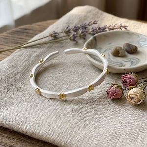 May include: A silver-tone bracelet with small gold-colored flower accents. The open cuff bracelet is displayed on a neutral-colored linen surface, with dried flowers and a small decorative dish in the background. Jewelry.