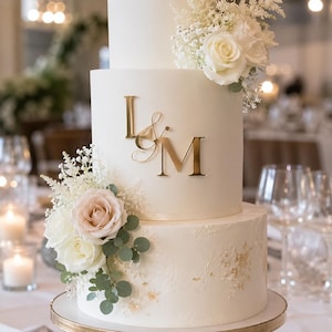 Può includere: Una torta nuziale bianca a più piani con glassa liscia, lettere monogramma L e M in metallo dorato e composizioni decorative di rose bianche, rose rosa pallido, gipsofila e foglie di eucalipto verde sui lati dei piani.