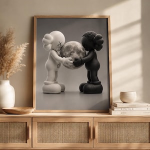 May include: A framed wall art print featuring a white figure and a black figure facing each other, both holding a textured gray sphere between them. The figures have rounded ears with X-shaped eyes and are depicted against a neutral gray background.